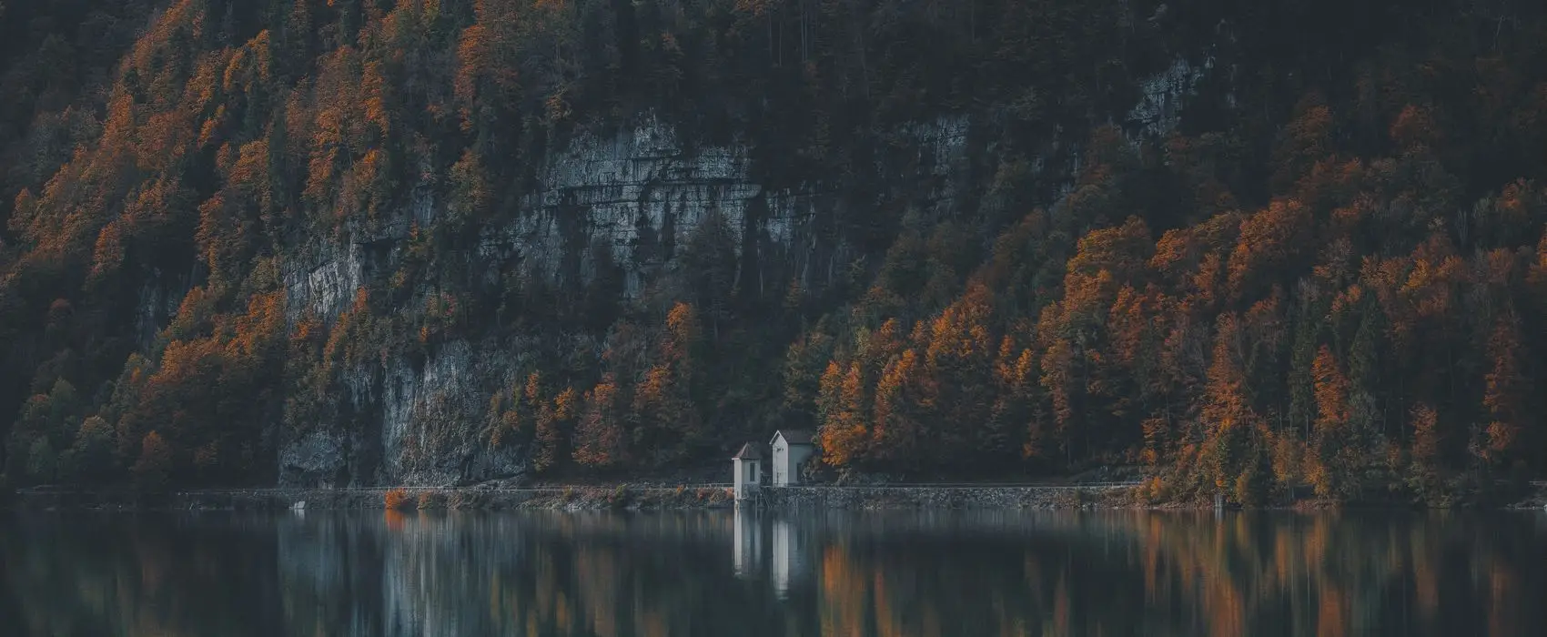 Ein ruhiger See mit einem kleinen Gebäude am Ufer, umgeben von herbstlichen Bäumen und einem steilen Felsen.