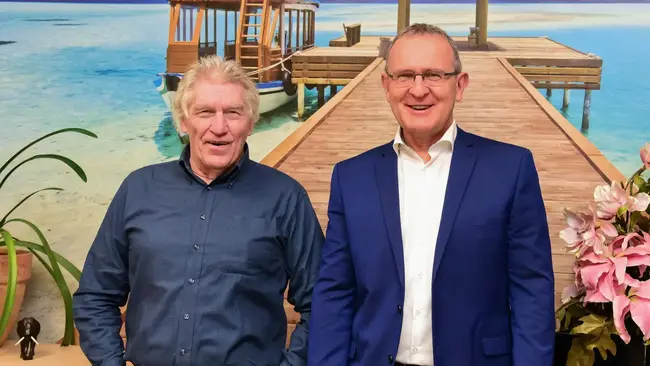 Zwei Männer stehen lächelnd vor einer Holzplattform am Wasser, umgeben von Pflanzen.